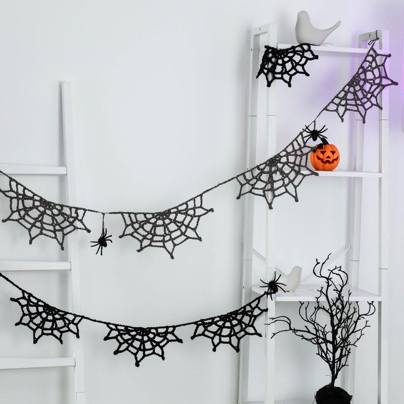 May include: A black crocheted spider web garland featuring multiple web segments connected by a string. Small black spider figures are attached to the webs. The garland is draped across a white ladder shelf, which also holds a small orange pumpkin.