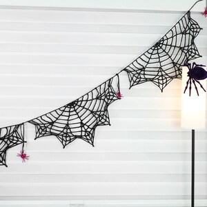 May include: A black crocheted spider web garland featuring multiple connected web segments. The garland is decorated with small, bright pink plastic spiders attached to the black yarn webbing at various points along the length.
