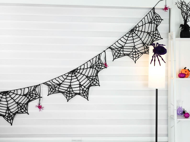 May include: A black crocheted spider web garland featuring multiple connected web segments. The garland is decorated with small, bright pink plastic spiders attached to the black yarn webbing at various points along the length.