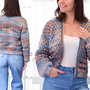 May include: A multi-colored, open-front crochet cardigan in shades of blue, tan, and brown. The handmade sweater has long sleeves and a cropped length. The open weave design is visible. The model is wearing blue jeans and a white top.