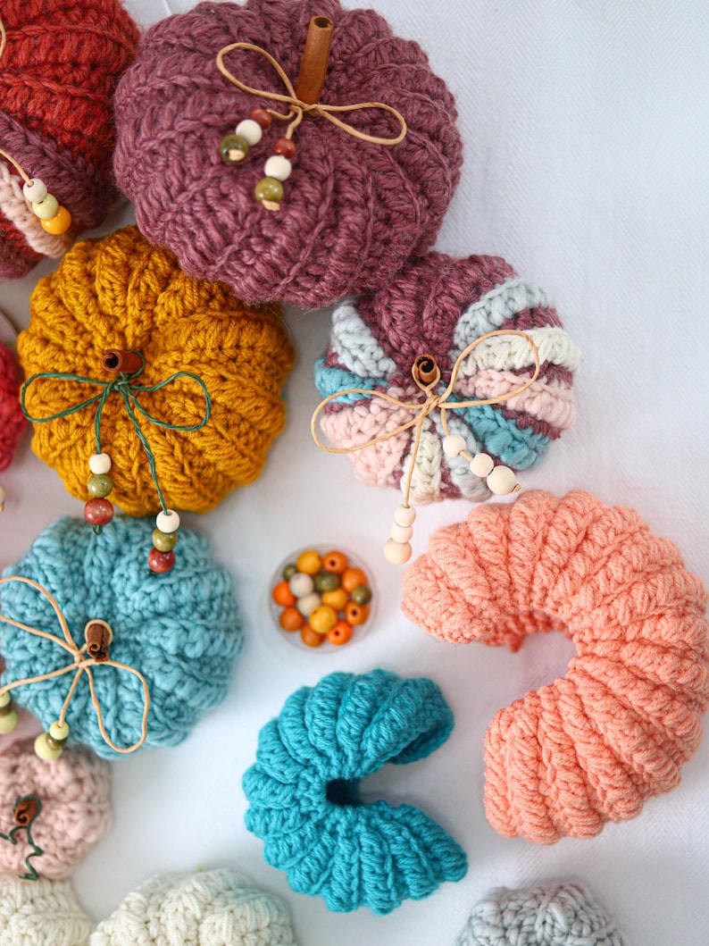 May include: A collection of crocheted pumpkins in various colors including plum, mustard yellow, teal, peach, and cream. Each pumpkin features a cinnamon stick stem, twine bow, and small wooden beads. A small container of loose wooden beads is included.