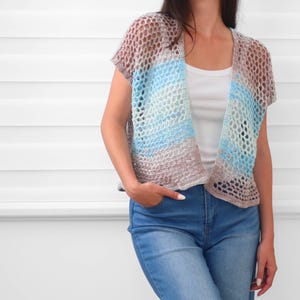 May include: A short-sleeved, open-front crochet cardigan featuring a loose, net-like stitch pattern. The garment displays a horizontal gradient of colors, transitioning from light brown at the top and bottom to shades of blue and white in the center.