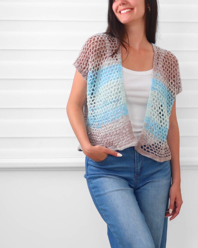 May include: A short-sleeved, open-front crochet cardigan featuring a loose, net-like stitch pattern. The garment displays a horizontal gradient of colors, transitioning from light brown at the top and bottom to shades of blue and white in the center.