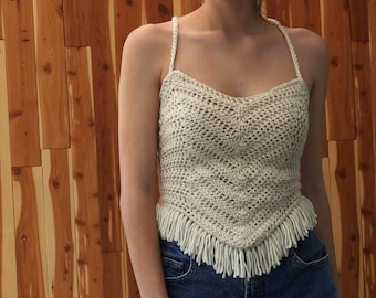 May include: A white crochet top with a fringe trim. The top has a scoop neck and thin straps.