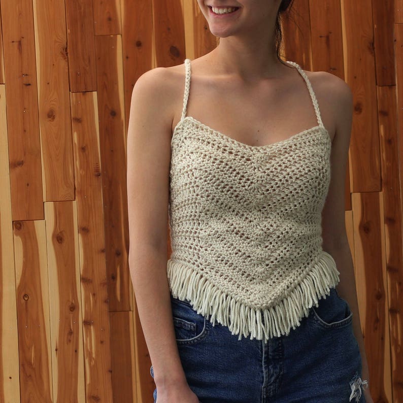 Puede incluir: Un top blanco de crochet con ribete de flecos. El top tiene un escote redondo y tirantes finos.