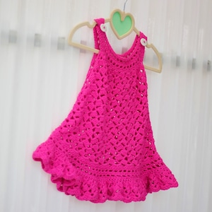 May include: A pink crocheted baby dress with a ruffled hem, hanging on a wooden hanger with a heart-shaped cutout.