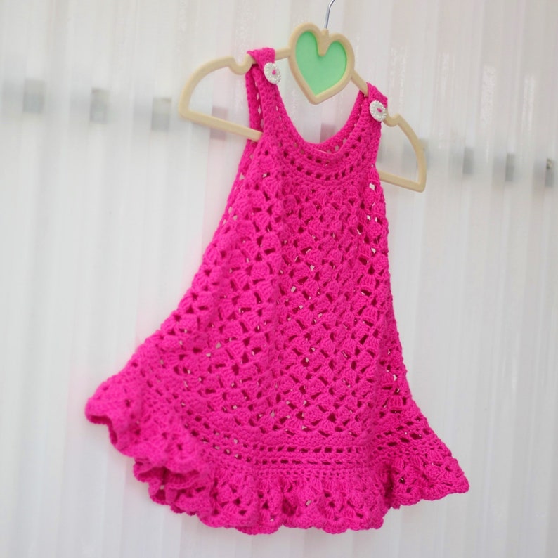 May include: A pink crocheted baby dress with a ruffled hem, hanging on a wooden hanger with a heart-shaped cutout.