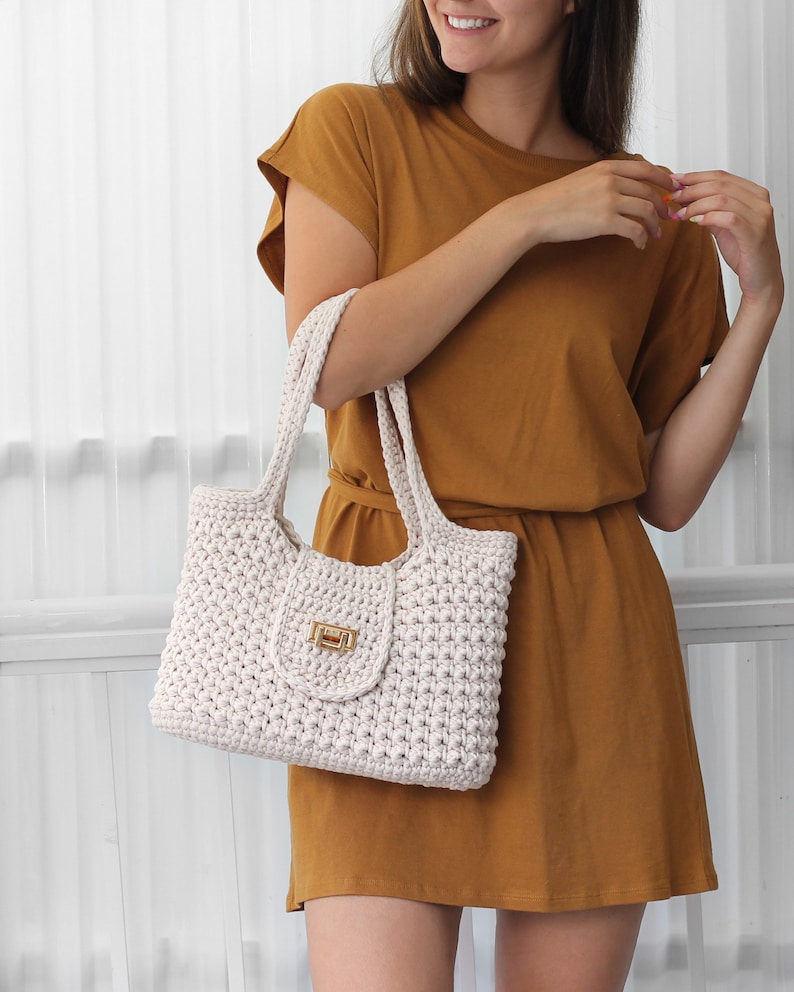 Puede incluir: Un bolso tote de crochet blanco con cierre de broche dorado. El bolso tiene un dise&ntilde;o simple y casual, perfecto para el uso diario.