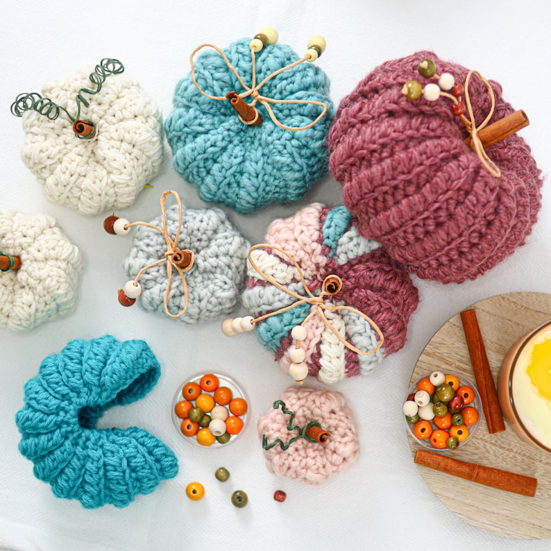 May include: A collection of crocheted pumpkins in various colors including white, teal, pink, and mauve. Each pumpkin features a cinnamon stick stem, some with twine and wooden beads. Loose wooden beads and cinnamon sticks are scattered nearby.
