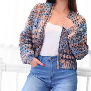 May include: A handmade crochet jacket in shades of blue, tan, and gray. The open-front cardigan features a textured stitch pattern and long sleeves. The jacket is paired with blue jeans.