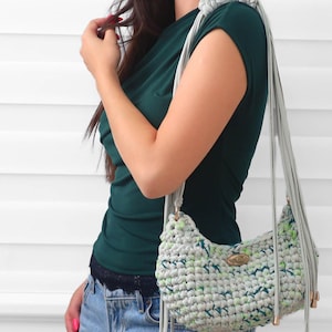 May include: A crescent-shaped crochet shoulder bag crafted from light gray yarn with subtle flecks of green and blue. The bag features a long, knotted gray strap and a small oval metallic brand tag on the front, finished with dangling metallic accents.