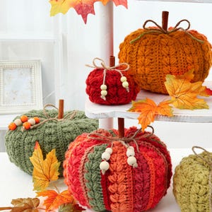 May include: A collection of crocheted pumpkins in orange, red, and green, each topped with a cinnamon stick stem, twine, and wooden beads. The pumpkins are arranged on a white tiered stand, accented with artificial autumn maple leaves.