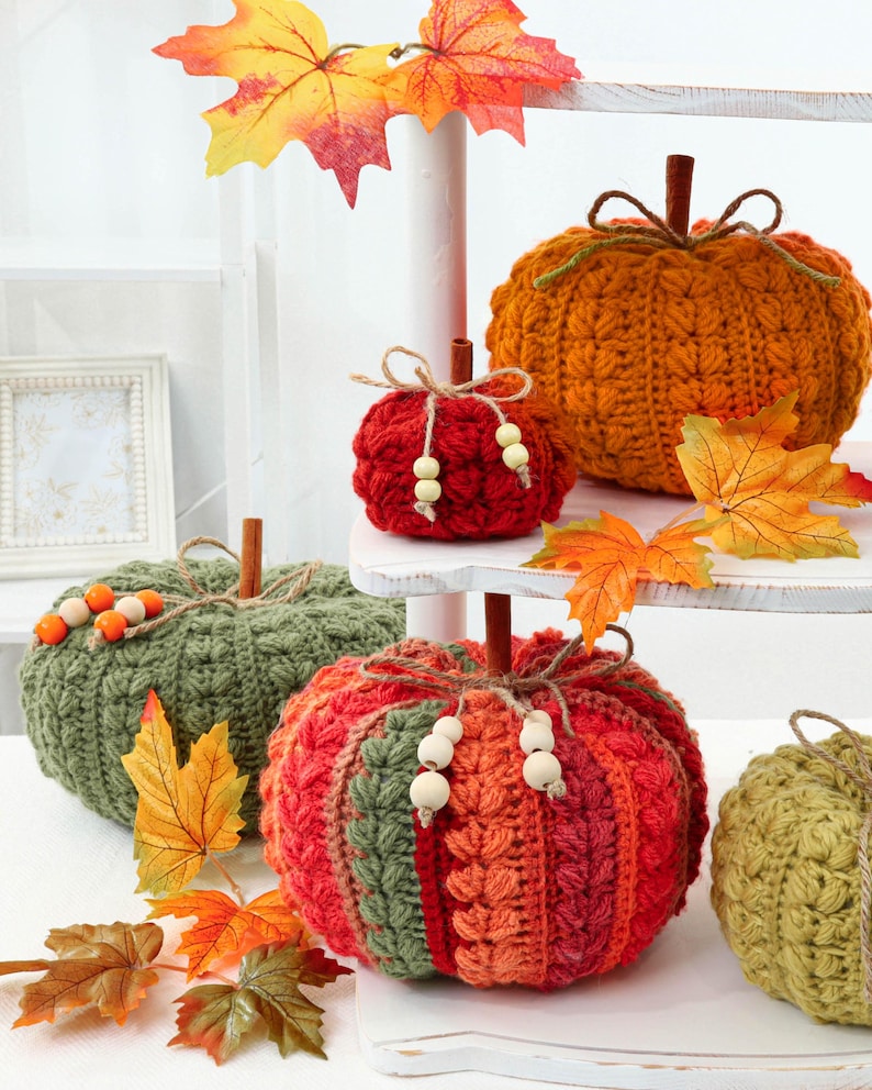 May include: A collection of crocheted pumpkins in orange, red, and green, each topped with a cinnamon stick stem, twine, and wooden beads. The pumpkins are arranged on a white tiered stand, accented with artificial autumn maple leaves.