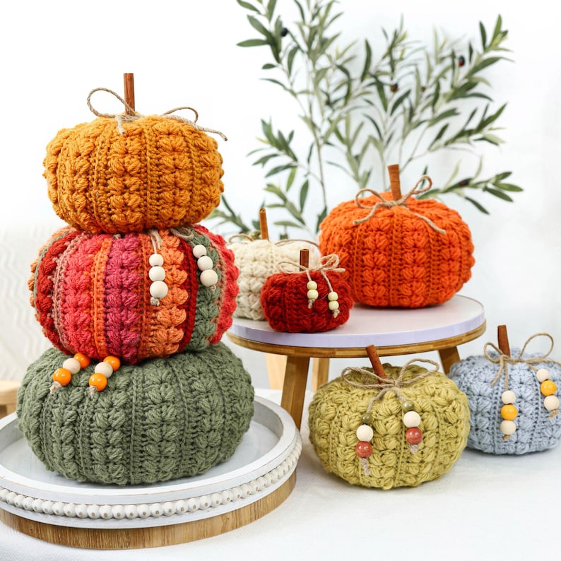 May include: A collection of handmade crochet pumpkins in various sizes and colors, including orange, red, green, cream, and light blue. Each pumpkin features a cinnamon stick stem, twine bow, and wooden beads. Some are stacked, others sit separately.
