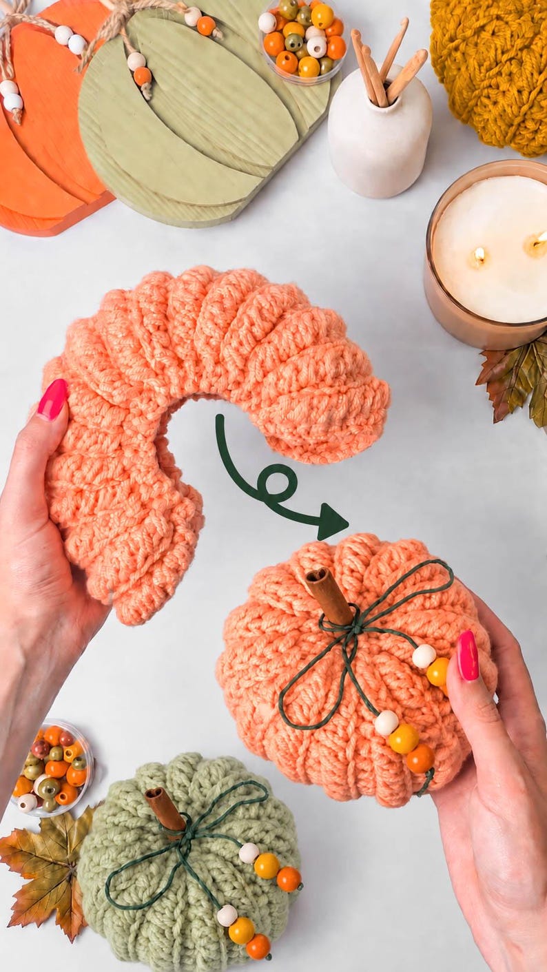 May include: A peach-colored crocheted pumpkin with a cinnamon stick stem and green twine tied with wooden beads. A dark green arrow points from a separate, curved peach-colored crocheted piece toward the assembled pumpkin.