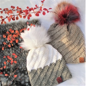 May include: Two crocheted hats with faux fur pom poms. The hats are beige and white with a chevron pattern. The pom poms are white and red and black.