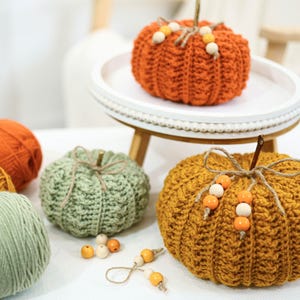 May include: A collection of handmade crochet pumpkins in various colors, including orange, green, and gold. Each pumpkin features a stem and decorative accents like wooden beads. Yarn skeins in coordinating colors are also present, suggesting a crafting theme.