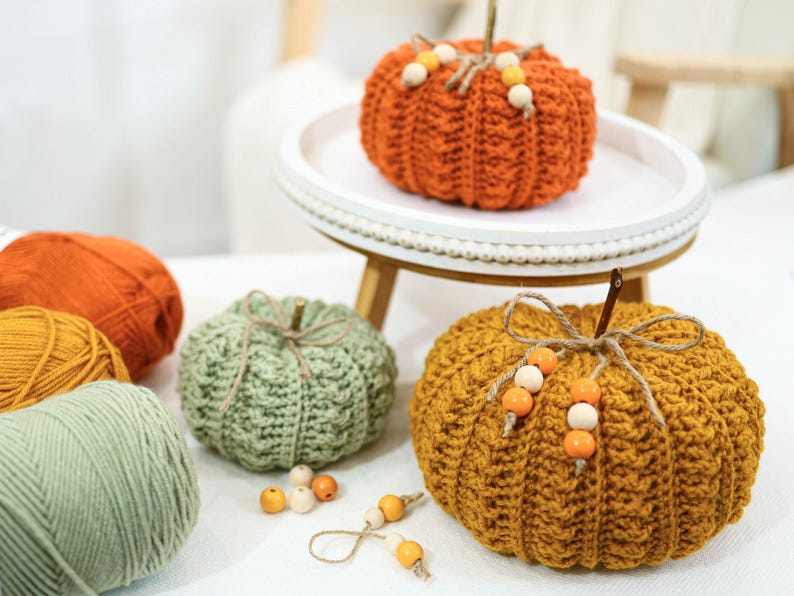 May include: A collection of handmade crochet pumpkins in various colors, including orange, green, and gold. Each pumpkin features a stem and decorative accents like wooden beads. Yarn skeins in coordinating colors are also present, suggesting a crafting theme.
