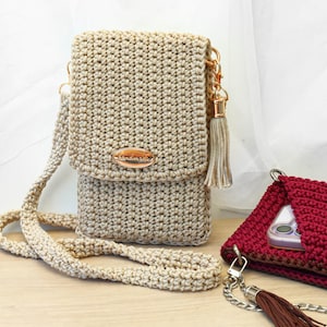 Könnte beinhalten: Beige gehäkelte Umhängetasche mit Klappe und langem Riemen. Die Tasche hat ein goldfarbenes Metallschild mit dem Wort "handmade" und einen passenden Quastenanhänger. Eine zweite rote Häkeltasche ist ebenfalls zu sehen.