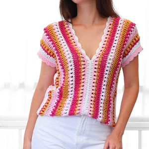 Pu&ograve; includere: Un top all'uncinetto colorato, a maniche corte e scollo a V. Il top presenta strisce verticali nei toni del rosa, bianco e giallo. Lo scollo e le maniche hanno un bordo smerlato. La modella indossa pantaloni bianchi.