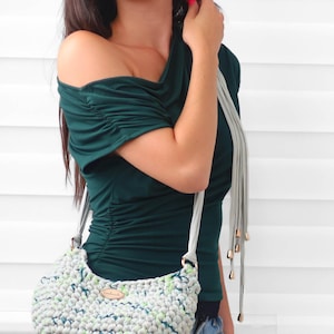 May include: A dark green short-sleeved top with gathered shoulder and side details, paired with a light gray crocheted handbag featuring a speckled pattern of blue and green, a light gray strap, and a small oval wooden tag on the front.