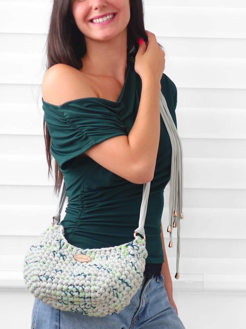 May include: A dark green short-sleeved top with gathered shoulder and side details, paired with a light gray crocheted handbag featuring a speckled pattern of blue and green, a light gray strap, and a small oval wooden tag on the front.