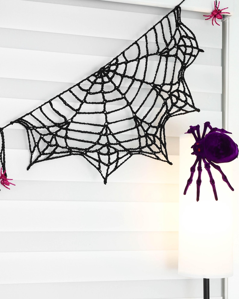 May include: A black crocheted spider web decoration featuring a series of interconnected yarn strands. The web is accented with small, bright pink glittery spiders attached to the edges and a large, dark purple spider positioned nearby.