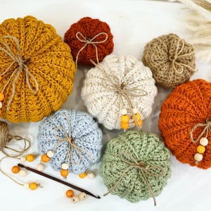 May include: A collection of handmade crochet pumpkins in various colors, including mustard yellow, orange, and light blue. Each pumpkin is adorned with twine and wooden beads, creating a rustic and autumnal aesthetic. Perfect for fall decor.