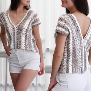 May include: A crocheted top with a v-neck and short sleeves. The top is made with a white, gray, and brown yarn and has a striped pattern. The top is shown on a model wearing white denim shorts.
