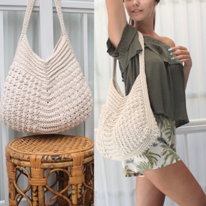 May include: Cream-colored crochet shoulder bag with a textured pattern. The bag has a wide, open top and a long strap. The bag is shown hanging and being worn. The bag is made of a soft, natural fiber.