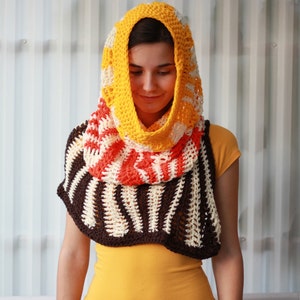 May include: A crocheted cowl with a hood in brown, white, orange, and yellow stripes. The cowl is being worn by a person with brown hair.