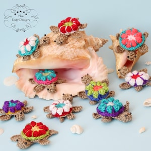 May include: A collection of crocheted turtle ornaments with colorful flower designs on their shells. The turtles are arranged around a large seashell.