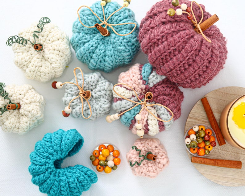 May include: A collection of crocheted pumpkins in various sizes and colors including white, teal, light blue, pink, and dark mauve. Each pumpkin features a cinnamon stick stem, twine, and wooden beads. Small bowls of colorful wooden beads are included.