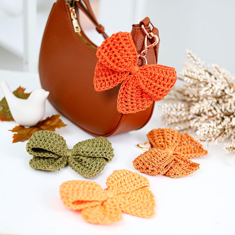 May include: A brown leather hobo bag with a gold zipper and a large orange crocheted bow keychain attached to the strap. Three additional crocheted bows in olive green, burnt orange, and light yellow are arranged nearby on the surface.
