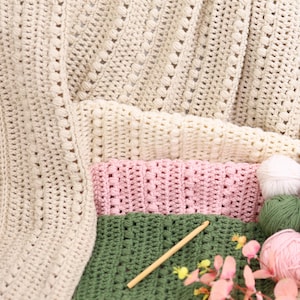 Pu&ograve; includere: Una collezione di articoli all'uncinetto fatti a mano in vari colori. L'immagine presenta coperte all'uncinetto color crema, rosa e verde e gomitoli di lana. Sono visibili anche un uncinetto di legno e fiori rosa.