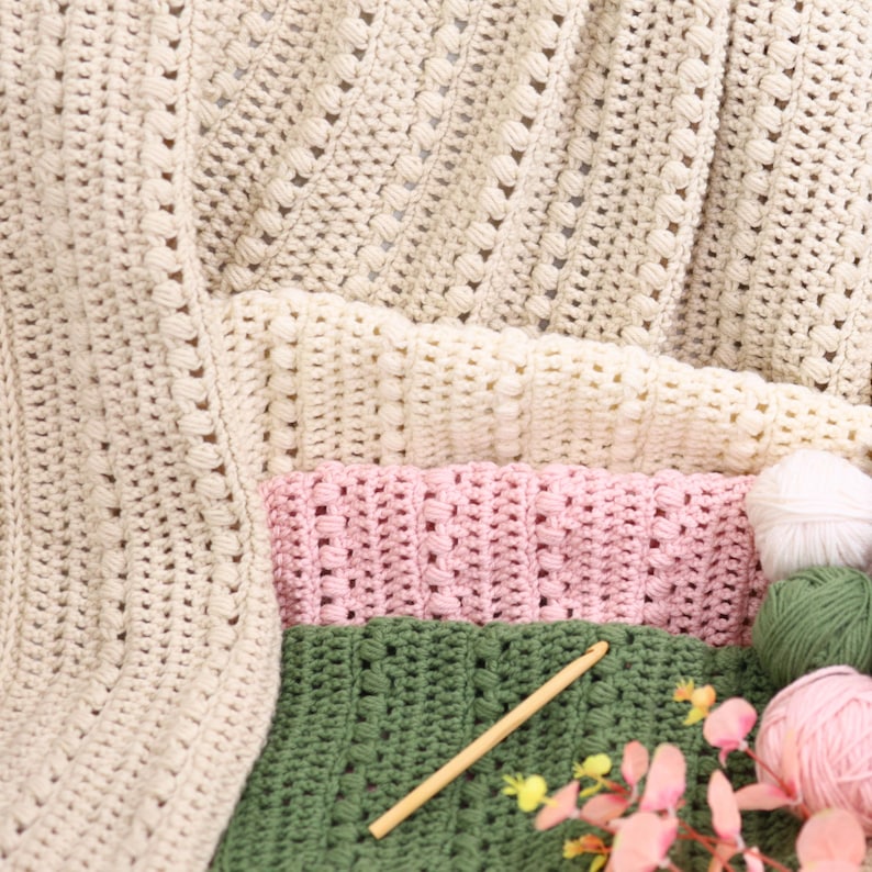 Pu&ograve; includere: Una collezione di articoli all'uncinetto fatti a mano in vari colori. L'immagine presenta coperte all'uncinetto color crema, rosa e verde e gomitoli di lana. Sono visibili anche un uncinetto di legno e fiori rosa.