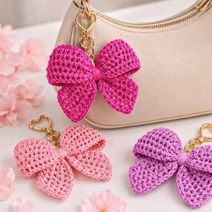 May include: Three crochet bow keychains in bright pink, light pink, and purple, each featuring a gold-toned heart-shaped lobster clasp. The bright pink bow is attached to a beige handbag, while the other two lie flat nearby.