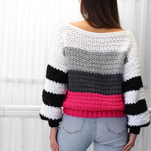 May include: A striped crochet sweater with a pink, gray, and white color scheme. The sweater has a loose fit and long sleeves.