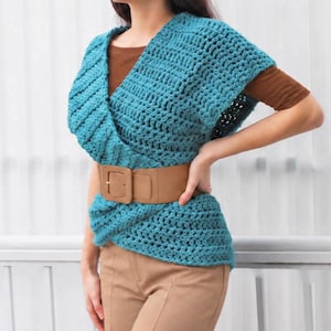 May include: A teal crochet wrap top with a V-neckline and short sleeves, worn over a brown short-sleeved shirt. The top is cinched at the waist with a wide tan belt featuring a large rectangular buckle, paired with tan trousers.