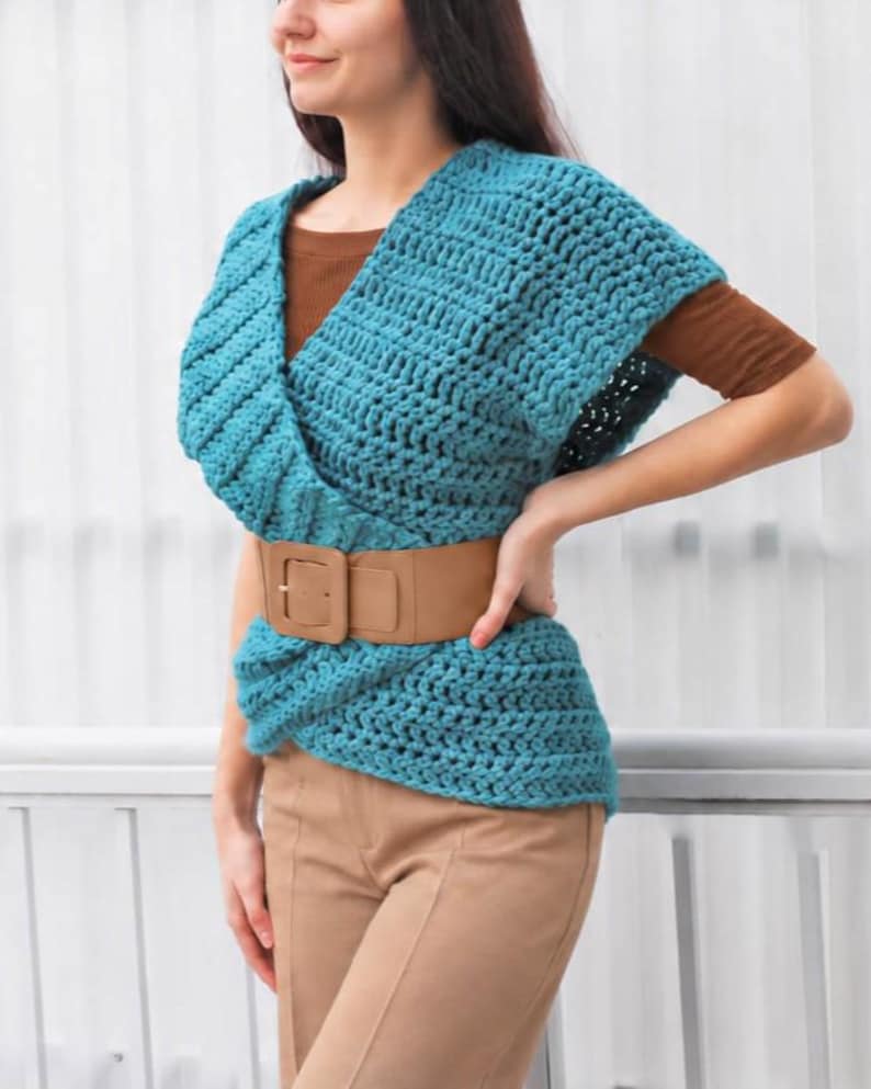 May include: A teal crochet wrap top with a V-neckline and short sleeves, worn over a brown short-sleeved shirt. The top is cinched at the waist with a wide tan belt featuring a large rectangular buckle, paired with tan trousers.
