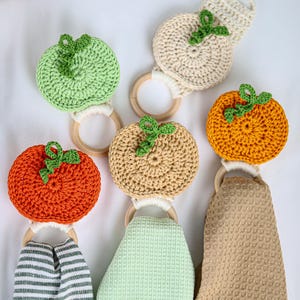 May include: A collection of five crochet fruit-shaped toppers attached to wooden rings, featuring orange, light green, and beige pumpkins and apples with green stems. Three of the rings are paired with hanging waffle-weave kitchen towels.