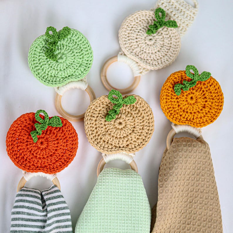 May include: A collection of five crochet fruit-shaped toppers attached to wooden rings, featuring orange, light green, and beige pumpkins and apples with green stems. Three of the rings are paired with hanging waffle-weave kitchen towels.