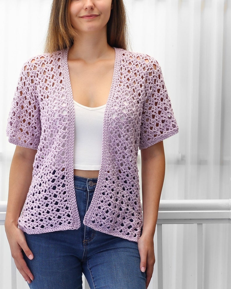 May include: A light purple crochet cardigan with a V-neck and short sleeves. The cardigan is made with a delicate open stitch pattern.