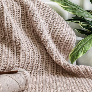 May include: A beige crocheted throw blanket with a textured stitch pattern. The blanket is folded and draped over a white surface with a green plant in the background. A ball of yarn and a crochet hook are in the foreground.