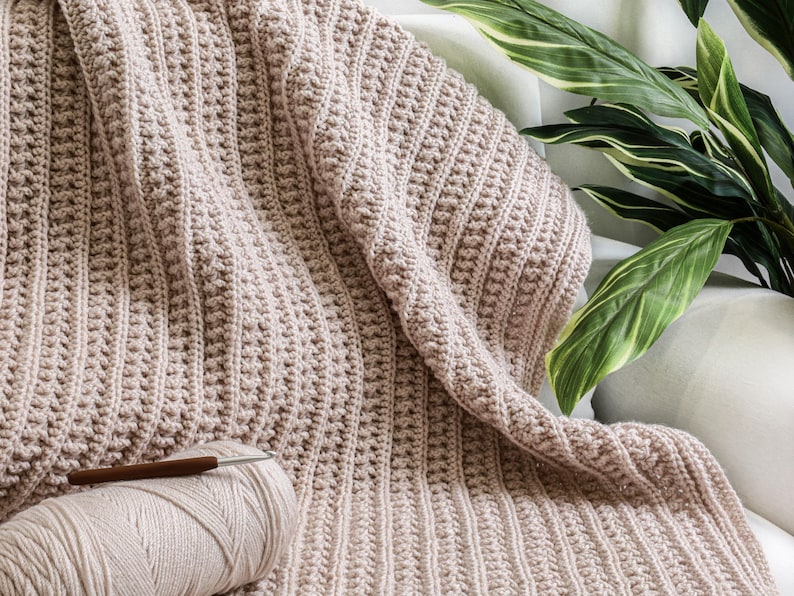 May include: A beige crocheted throw blanket with a textured stitch pattern. The blanket is folded and draped over a white surface with a green plant in the background. A ball of yarn and a crochet hook are in the foreground.