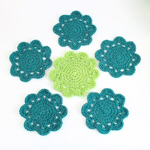 Op de afbeelding: Zes gehaakte onderzetters in teal en groen. De onderzetters zijn rond en hebben een gegolfde rand. Ze zijn perfect om een ​​toets van handgemaakte charme aan uw huisdecor toe te voegen.