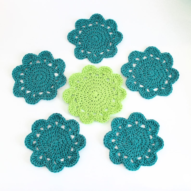 Op de afbeelding: Zes gehaakte onderzetters in teal en groen. De onderzetters zijn rond en hebben een gegolfde rand. Ze zijn perfect om een ​​toets van handgemaakte charme aan uw huisdecor toe te voegen.