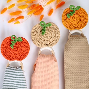 May include: Three hanging kitchen towels, each featuring a crocheted pumpkin topper in orange or beige with a green stem, attached to a wooden ring. The towels have different textures, including a gray and white stripe and solid waffle weaves.