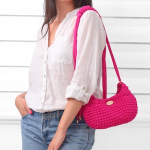 May include: A vibrant hot pink crochet shoulder bag featuring a textured, knotted stitch pattern. The bag includes a matching pink strap with decorative fringe accents and a small, oval-shaped metallic brand tag centered on the front exterior.