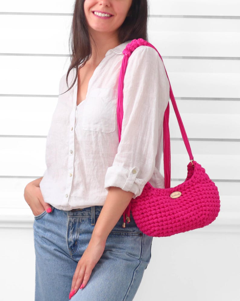 May include: A vibrant hot pink crochet shoulder bag featuring a textured, knotted stitch pattern. The bag includes a matching pink strap with decorative fringe accents and a small, oval-shaped metallic brand tag centered on the front exterior.